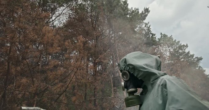 Man testing the branches of pine trees for contamination in a WWII Hasmat suit and gas mask as smoke or toxic vapor billow around him.