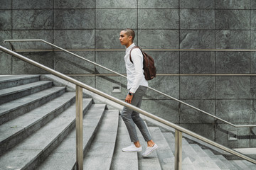 Young man dressed in casual clothes and backpack in the city center is going up the stairs -...