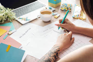 cropped view of woman sitting behind wooden table with laptop and stationery, writing in paper...