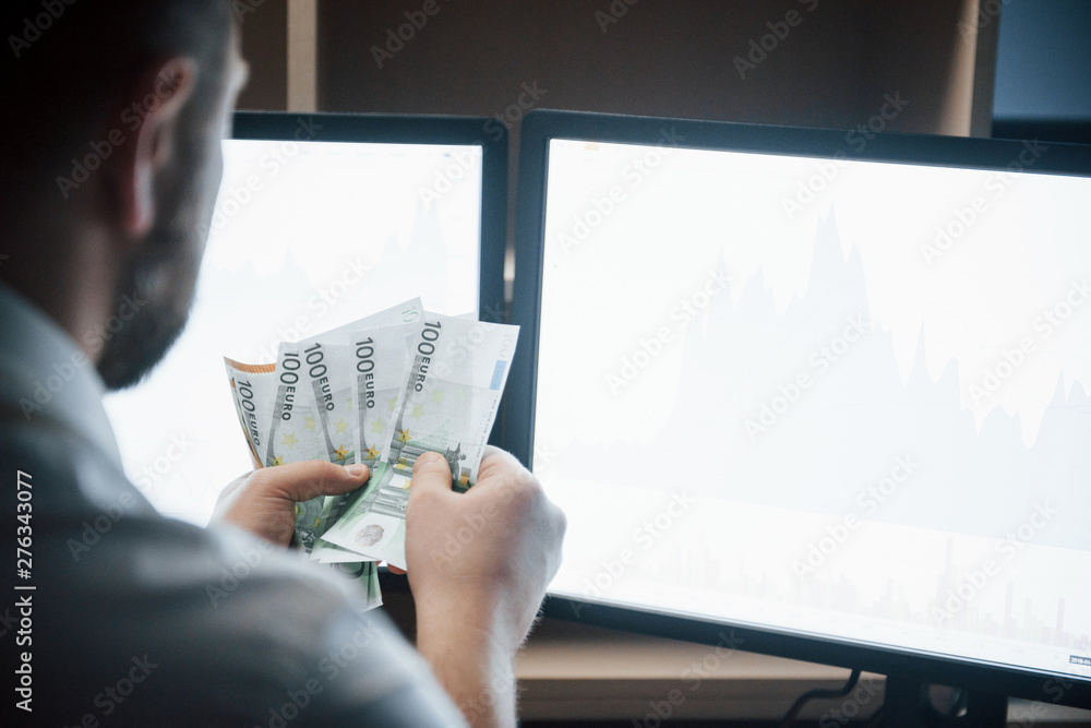 Poster view from behind. bearded man in white shirt holds earned money in the office with multiple computer