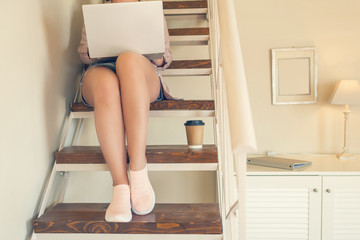 Girl student in shorts sits on the steps with a laptop and paper cup of coffee in a cafe in the home style