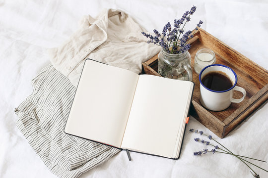 French Summer Still Life. Feminine Lifestyle Composition. Cup Of Coffee, Lavender Flowers Bouquet, Candle On Wooden Tray. Linen Shirt Nad Trousers On Bed. Blank Notebook Mockup Scene. Top View.