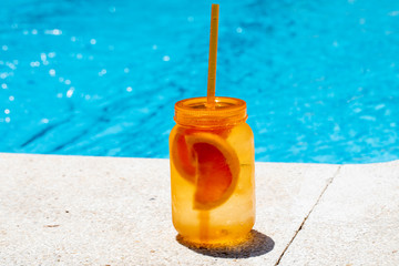 Summer cold drink. Orange mason jar on the bord of a swimming pool with bright blue water