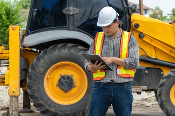 Engineering wearing a white safety helmet standing In front of the backhoe And are using tablet for...