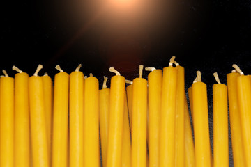 Many natural thin yellow wax candles lie on black background.