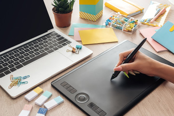 cropped view of woman working with drawing tablet, sitting behind wooden table with stationery and laptop