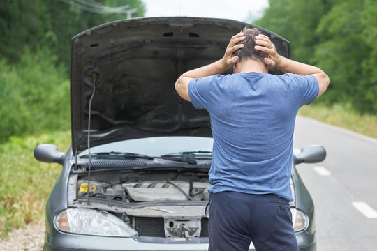 Middle-aged worried man stands with his hands on the head near his broken car with raised hood on the rural road