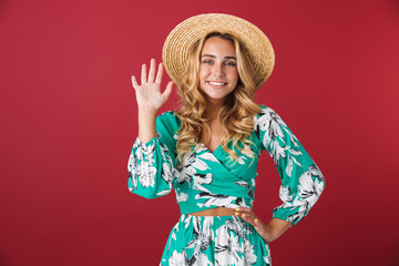 Smiling happy young blonde girl in bright blue dress posing isolated over red wall background waving to you.