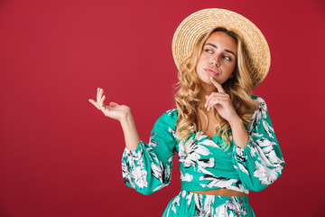 Thinking thoughtful cute young blonde girl in bright blue dress posing isolated over red wall background looking aside wearing hat showing copyspace.