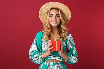 Cute young blonde girl in bright blue dress posing isolated over red wall background holding present box.