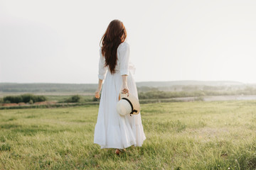 Beautiful carefree long hair girl in white clothes and straw hat enjoys life in nature field at sunset. Sensitivity to nature concept
