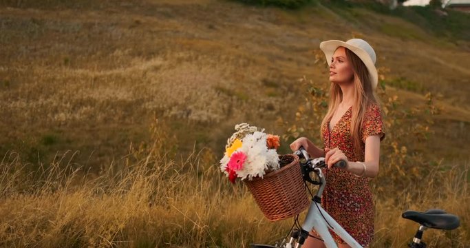 Young smiling blonde in hat and dress walking in dress with bike and flowers in basket.