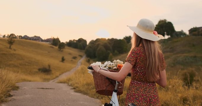 Back plan: Beautiful blonde in dress and retro bike walking on the road in the summer field.