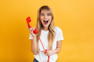 Happy young surprised blonde woman posing isolated over yellow wall background talking by retro red telephone.