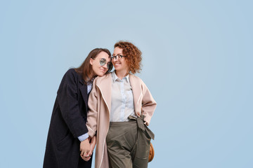 Portrait of laughing girls with hands on shoulders and glasses. Young happy girlfriends over blue isolated background. Smiling young female in casual coat having fun. Look at camera and each other.