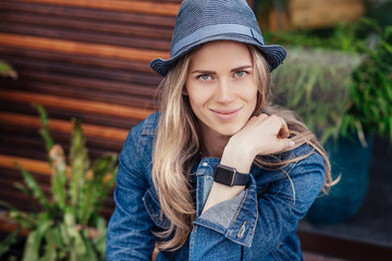 Portrait of pretty young female student in casual clothes with smart watches posing near a wooden cafe on a sunny warm summer day. The concept of planning and reminders. Copyspace