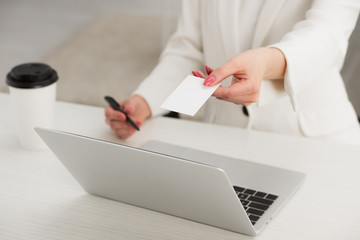cropped view of businesswoman holding blank business card and pen while sitting at desk near laptop