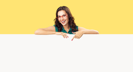 Happy smiling beautiful young woman in green clothing and red glasses, pointing blank white bill...