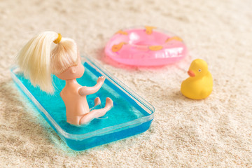 Baby girl doll sitting in kids swimming pool next to inflatable pool float and rubber duck on the beach minimal creative summer vacation concept.