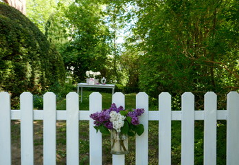 White garden fence in front of an idyllic garden. On the fence hangs a glass vase with a bouquet of white and purple blooming lilac.