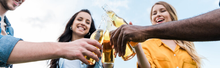 panoramic shot of cheerful multicultural men and women clinking bottles with beer
