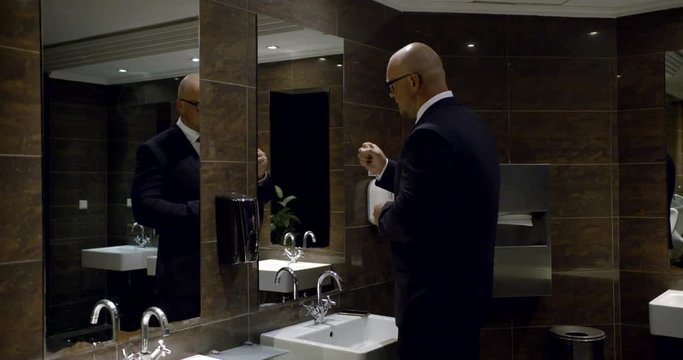 In the men's room, a bald man in glasses and a business suit, standing near the sink, threatens himself in the mirror with his fist, washes his hands, then takes a paper towel and wipes their hands