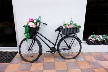Retro bicycle as a flowerbed decoration outside element. Black aged bike stays against the wall with two garden baskets with beauty flowers. Art decor object