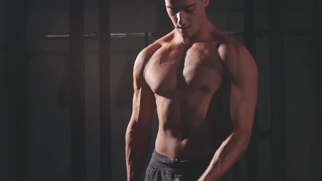 Shirtless white man in sweatpants starting exercise with dumbbell weight in dark gym. Fitness motivation and muscle training concept.