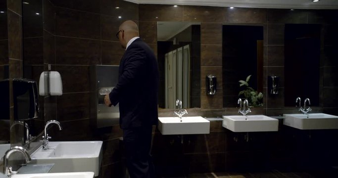 In the men's room, a bald man in glasses and a business suit stands by the sink and washes his hands, shakes them off at the end, then takes a paper towel and wipes their hands