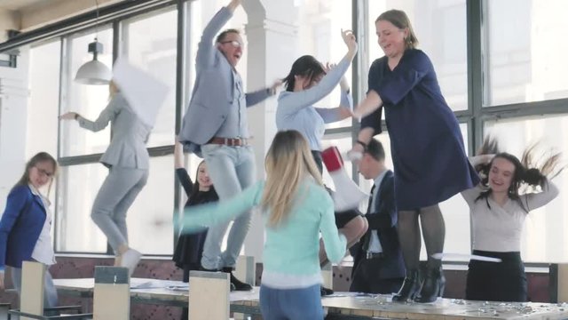 A business team cheerfully dances on a table in an office space, sings into a megaphone, throws documents and celebrates success. Office holiday. Corporate