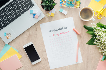top view of 100 days check list, stationery and laptop with smartphone on wooden table