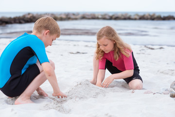 Two children in neoprene swimsuits playing on the beach with sand