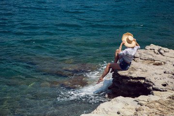 woman in a hat and shirt sit on a stone by the sea