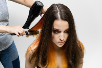 Fototapeta premium Photo portrait of the work of the master hairdresser makes a fashionable hairstyle with a hair dryer and a hairbrush in the studio on a white background. The brunette model has colored hair.