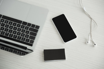 top view of laptop, smartphone with blank screen, earphones and stack of black empty business cards on office table