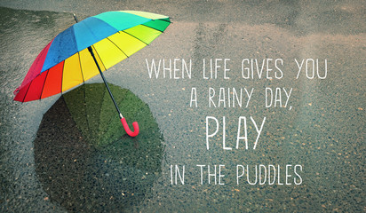 when life gives you a rainy day, play in the puddles - inspiration quote on autumn abstract blurred defocused background. rainbow umbrella in puddle, rainy weather season. soft selective focus