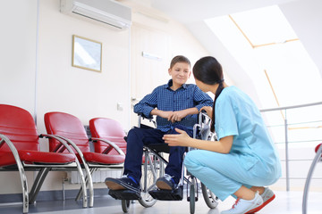 Young female doctor taking care of little boy in wheelchair indoors