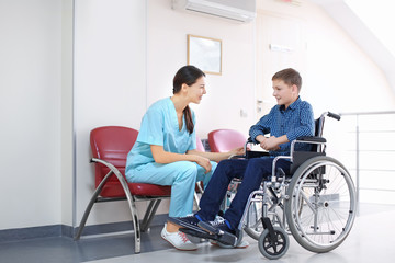 Young female doctor taking care of little boy in wheelchair indoors