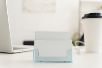 card holder with blank business cards near paper cup and laptop on table in office