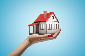 Side closeup of woman's hand facing up and holding small one-storey house on light blue gradient background.
