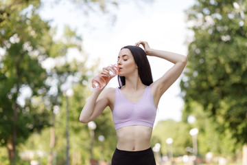 Beautiful slim brunette woman drinking water from bottle after running at the  morning park to stay hydrated. Female fitness model working out outdoor. Concept of healthy lifestyle.