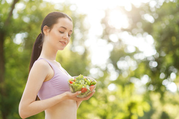 Beautiful slim brunette eating salad over green natural background. Female fitness model at the park. Woman hands holding fresh summer salad with raw vegetables cucumbers tomatoes lettuce in bowl.