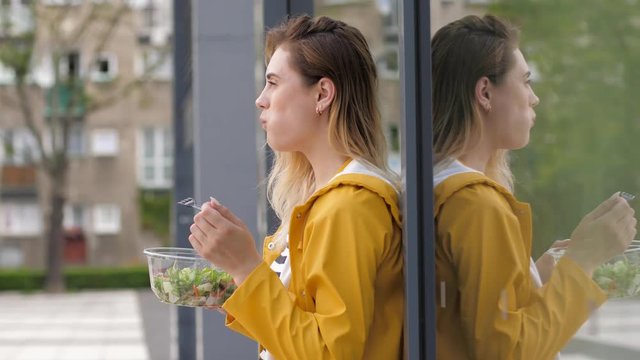 Beautiful young girl eat healthy vegetable salad from plastic bowl staying alone in yellow coat leaning a glass wall surface