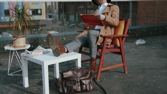 Attractive stylish mature man having a nice break on the wide terrace above the bautiful city panorama. Guy in hat typing on a computer laptop sitting in chair working online outdoors.