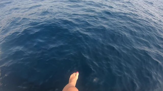 Caucasian woman filming her legs and feet hanging over the deep ocean of malta on a boat while she is travelling to her next holiday adventure.