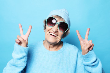 Funny old lady wearing blue sweater, hat and sunglasses showing victory sign. Isolated on blue background.