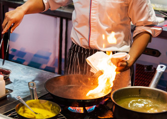 Chef cooking with flame in a frying pan on a kitchen stove.