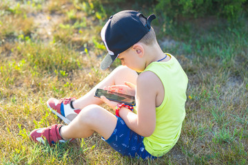 Little boy sits in the park on the ground and looks into the smartphone