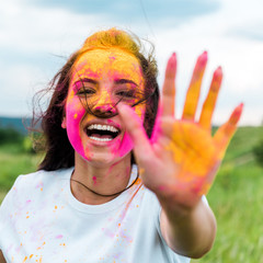 selective focus of cheerful woman with pink and yellow holi paint on face and hands