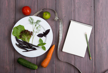 Healthy diet plan concept - note pad for recipe, measure tape, white plate with leaf of salad, cucumber, tomato and other vegetable